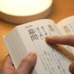 開かれた国語辞典の「幕間」の項目を指差す日本人の手。正しい読み方「まくあい」が強調されている。