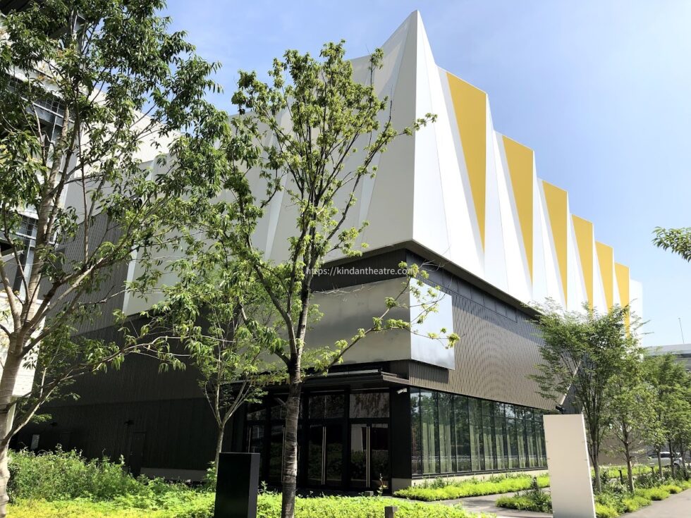 黄色と白の幾何学的な外壁デザインが特徴的な有明四季劇場の外観と周辺の緑